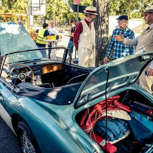 Eine Gruppe von Männern in stilvollen Outfits steht um ein klassisches Auto. Der Kofferraum des Fahrzeugs ist geöffnet und zeigt Gepäck.
