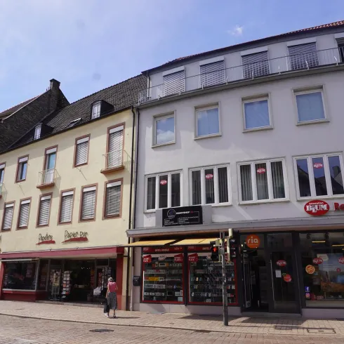 Eine Straße mit verschiedenen Gebäuden und Geschäften. Die Fassaden sind bunt und gut erhalten, bei schönem Wetter.