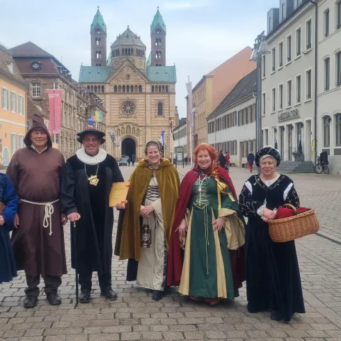 Sechs Personen in historischer Kleidung stehen auf einem Platz. Im Hintergrund sind alte Gebäude und ein Kirchturm zu sehen.