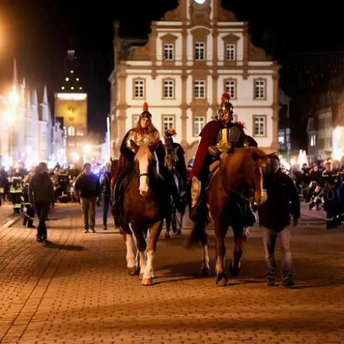 Zwei Reiter in historischen Kostümen reiten auf Pferden durch eine belebte Straße bei Nacht. Im Hintergrund sind Menschen und beleuchtete Gebäude zu sehen.