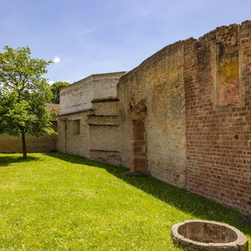 Eine grüne Wiese umgeben von alten Backsteinmauern. Im Hintergrund steht ein Baum und ein runder Brunnen ist sichtbar.