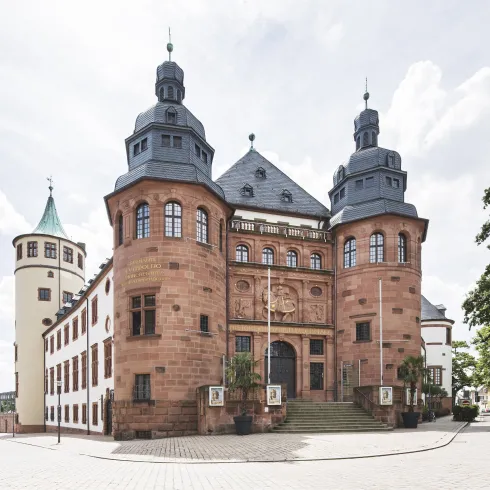 Ein historisches Gebäude mit schönem Fassadendesign und Türmen. Die Architektur zeigt traditionelle Elemente und ist von einem öffentlichen Platz umgeben.
