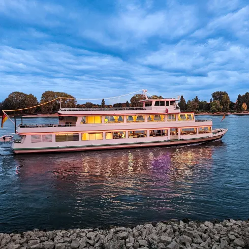 Ein Boot fährt gemütlich auf einem Fluss unter einem bewölkten Himmel. Die Lichter des Bootes spiegeln sich im Wasser.