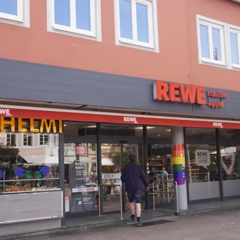 Ein REWE-Supermarkt mit einer einladenden Fassade. Die Eingangstür ist gut sichtbar und es gibt bunte Dekorationen.