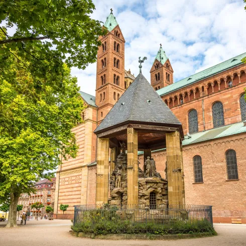Eine beeindruckende Kirche mit zwei Türmen und einer kunstvollen Statue im Vordergrund. Umgeben von Bäumen und einem einladenden Platz.