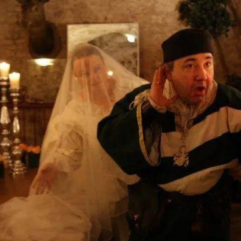 Ein kostümierter Mann hält eine Kerze und scheint zu reden, während eine Frau im Brautkleid hinter ihm steht. Die Szene spielt in einem stimmungsvollen Raum mit Kerzenlicht und historischem Ambiente.