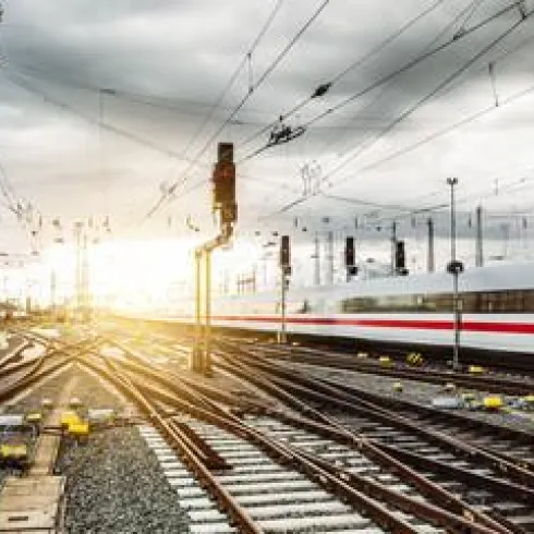 Ein Eisenbahnnetz mit vielen Gleisen und einem vorbeifahrenden Zug. Im Hintergrund ist ein Sonnenuntergang hinter grauen Wolken sichtbar.