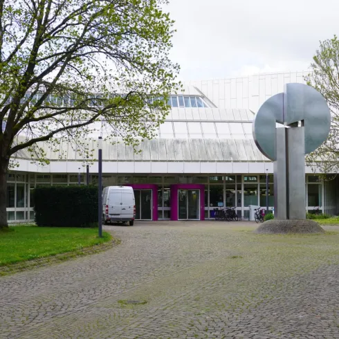 Ein modernes Gebäude mit Glasfenstern und einem skulpturalen Kunstwerk im Vordergrund. Im Hintergrund sind Bäume und ein gepflasterter Weg zu sehen.
