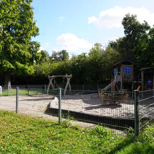 Ein Spielplatz mit Sandkasten, Schaukeln und einem Spielhaus. Umgeben von Bäumen und einem Zaun.
