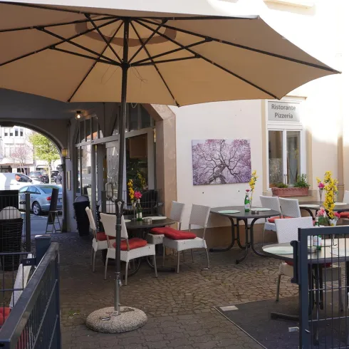 Ein gemütlicher Außenbereich eines Cafés mit Tischen und Stühlen. Ein großer Sonnenschirm bietet Schatten und farbige Blumen dekorieren die Tische.