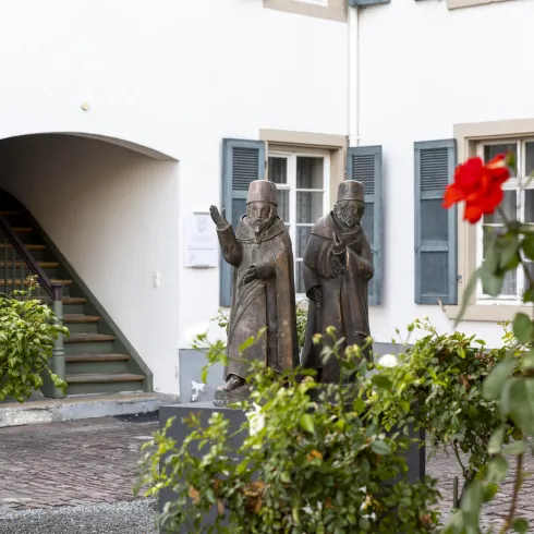 Zwei bronzene Statuen stehen in einem Innenhof mit grünen Pflanzen. Im Vordergrund blüht eine rote Rose.