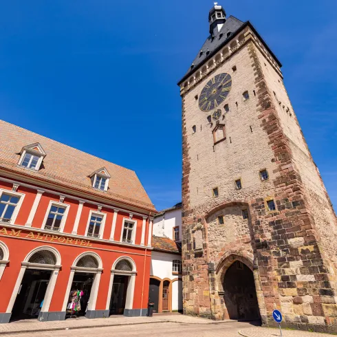 Ein historischer Turm mit einer Uhr in einer Stadt. Im Vordergrund sind schöne Gebäude und ein strahlend blauer Himmel zu sehen.