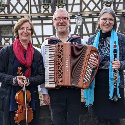 Drei Musiker stehen lächelnd zusammen. Sie halten verschiedene Musikinstrumente: eine Violine, ein Akkordeon und ein Klarinette.