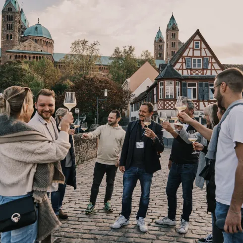 Eine Gruppe von Menschen feiert auf einer Brücke mit Gläsern in der Hand. Im Hintergrund sind historische Gebäude und Kirchen zu sehen.