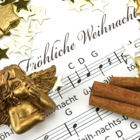 Ein goldener Engel sitzt auf Noten mit Weihnachtsliedern. Daneben sind Zimtstangen und goldene Sterne verstreut.