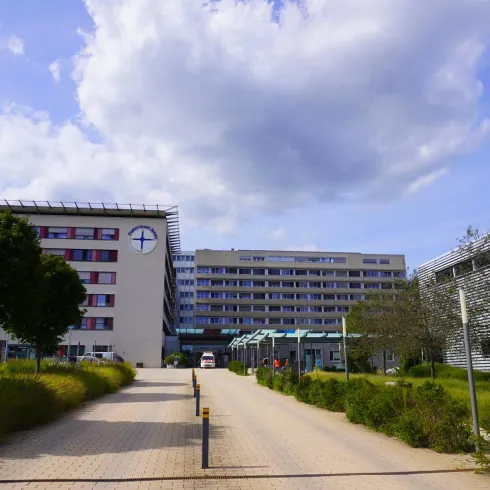 Ein modernes Krankenhausgebäude mit einer gepflasterten Zufahrt. Umgeben von grünen Flächen und Bäumen unter einem klaren Himmel.
