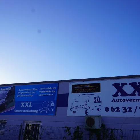 Ein Gebäude mit großen Werbeschildern für eine Autovermietung namens "XXL". Die Anzeige zeigt verschiedene Dienstleistungen und Kontaktinformationen.
