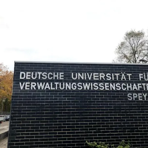 Ein Gebäude mit dem Schriftzug „Deutsche Universität für Verwaltungswissenschaften Speyer“. Im Hintergrund sind Bäume und eine Straße sichtbar.