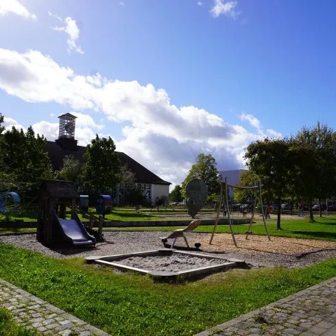 Ein Spielplatz in einem Park mit Rutschen und Schaukeln. Im Hintergrund sind Bäume und ein klarer blauer Himmel zu sehen.