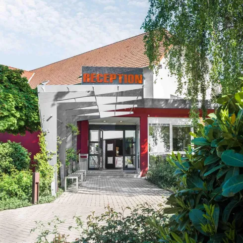 Ein Empfangsbereich mit großen Fenstern und einem modernen Eingang. Umgeben von üppigem Grün und Pflanzen.
