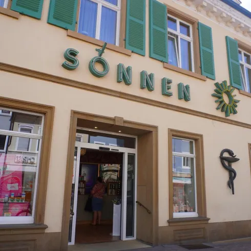 Eine Apotheke mit großem Eingang und grünem Fensterladen. Der Schriftzug „SÖNNEN APOTHEKE“ und ein Sonnensymbol sind deutlich sichtbar.