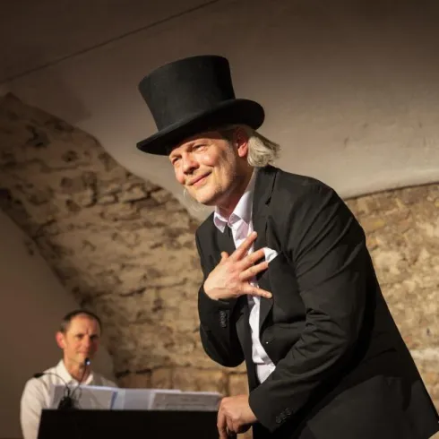 Ein Mann trägt einen Zylinder und einen Anzug, während er auf einer Bühne performt. Im Hintergrund sitzt ein Pianist und spielt Musik.