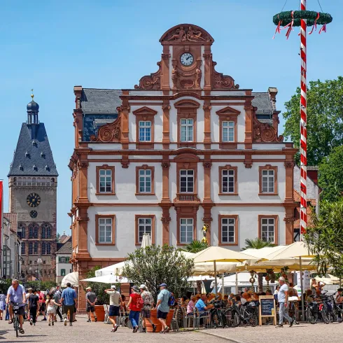 Ein belebter Platz mit schönen historischen Gebäuden und vielen Menschen. Im Hintergrund sind Bäume und Stände mit Sonnenschirmen zu sehen.