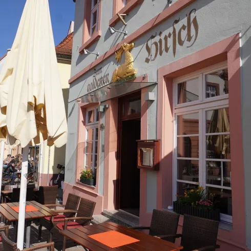Ein charmantes Gasthaus mit dem Namen "Hirsch" und einem einladenden Außenbereich. Tische und Stühle stehen bereit, während Sonnenschirme Schatten spenden.