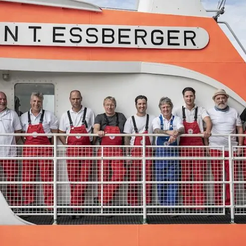 Eine Gruppe von Männern in roten Overalls steht auf einem Schiff mit der Aufschrift "JOHN T. ESSBERGER". Sie lächeln und posieren gemeinsam auf dem Vorderdeck.