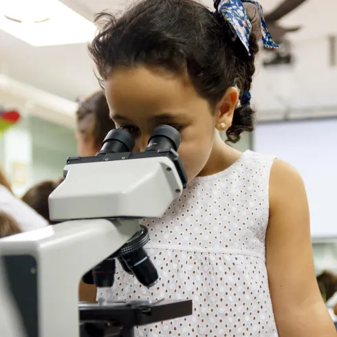 Ein kleines Mädchen schaut durch ein Mikroskop. Im Hintergrund sind weitere Personen und ein hell erleuchteter Raum zu sehen.