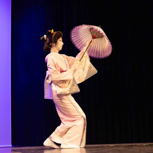Eine Tänzerin in einem traditionellen Kimono führt eine Darbietung mit einem Fächer auf. Sie bewegt sich elegant auf der Bühne vor einem dunklen Hintergrund.