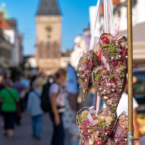 Hängende herzförmige Dekorationen in einer belebten Marktstraße. Im Hintergrund ist ein historischer Turm und viele Menschen zu sehen.