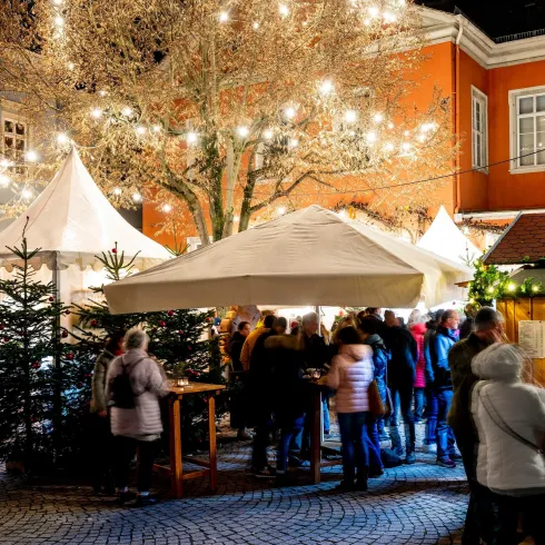 Ein festlicher Markt bei Nacht mit beleuchteten Ständen und geschmückten Bäumen. Viele Menschen genießen die Atmosphäre und die Angebote.