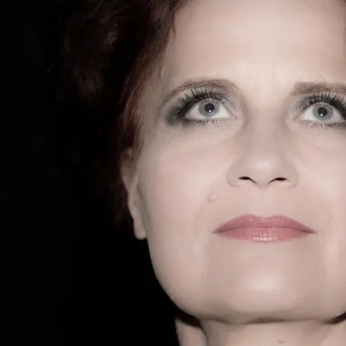 Eine Frau mit rotem Haar und intensiven Augen blickt nach oben. Ihr Gesicht ist sanft beleuchtet und wirkt geheimnisvoll.