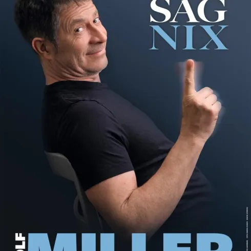Ein lächelnder Mann in einem schwarzen T-Shirt sitzt auf einem Stuhl und zeigt mit dem Finger nach oben. Der Text "ICH SAG NIX" und "ROLF MILLER" ist prominent im Bild zu sehen.