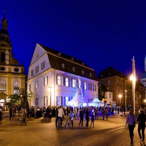 Eine lebhafte Abendveranstaltung auf einem Platz mit mehreren Gebäuden im Hintergrund. Die Szene ist mit vielen Menschen und festlicher Beleuchtung belebt.