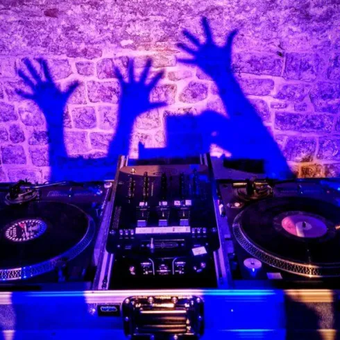 Eine DJ-Konsole mit Schallplatten und einem Mischpult. Die bunten Schatten von Händen sind an der Wand zu sehen.