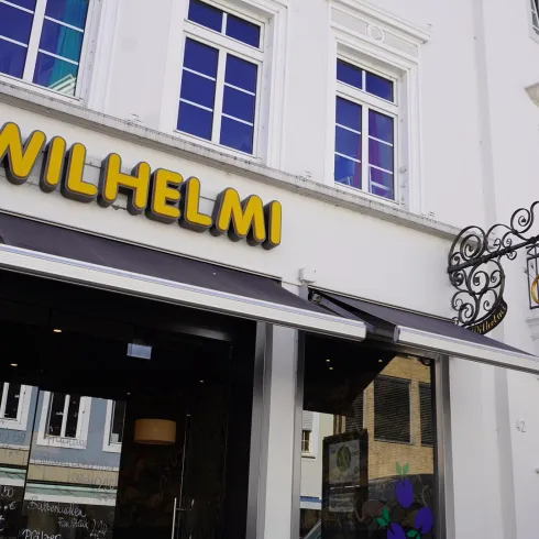 Ein Geschäft mit dem Namen "WILHELMI" an einer weißen Wand. Die Fassade hat große Fenster und einen eleganten, geschwungenen Balkon.