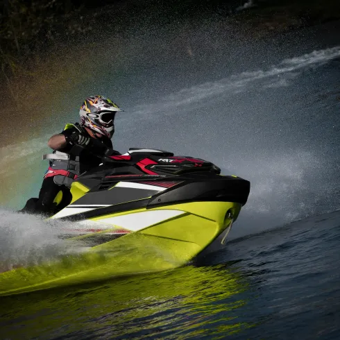 Ein Fahrer auf einem Jetski fährt durch das Wasser. Spritzwasser und ein Regenbogen sind im Hintergrund sichtbar.