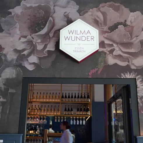 Ein gemütlicher Restaurant-Eingang mit einer eleganten Wandgestaltung aus Blumenmotiven. Über dem Eingang hängt ein Schild mit dem Namen "WILMA WUNDER".