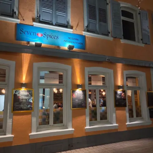 Ein einladendes Restaurant mit orangen Wänden und vielen Fenstern. Menschen stehen im Freien und genießen die abendliche Atmosphäre.