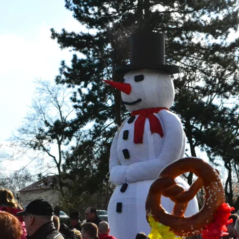 Eine große Schneemann-Figur mit einem Hut und einer Karottennase steht inmitten einer Menschenmenge. Im Vordergrund ist eine Brezel zu sehen.