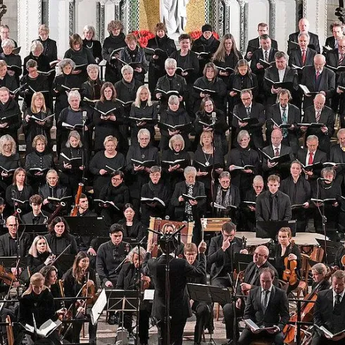 Ein großer Chor mit vielen Sängern steht in einer Kirche. Vor ihnen spielt ein Orchester auf der Bühne.