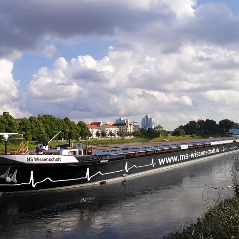 Ein Schiff mit dem Namen "MS Wissenschaft" fährt auf einem Fluss. Im Hintergrund sind Bäume und einige Gebäude zu sehen.