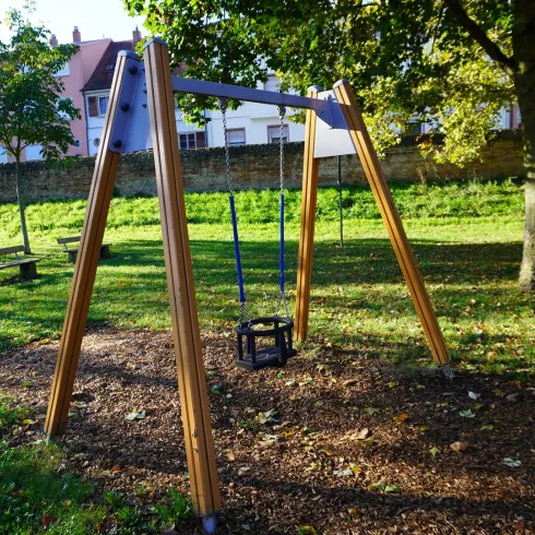 Eine Schaukel steht in einem ruhigen Park, umgeben von grünen Bäumen. Im Hintergrund ist eine Bank und einige Häuser sichtbar.