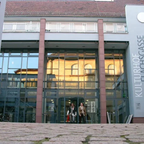 Ein modernes Gebäude mit Glasfassade und dem Schriftzug "Kulturhof Flachsgasse". Vor dem Eingang sind Menschen zu sehen, die das Gebäude betreten.