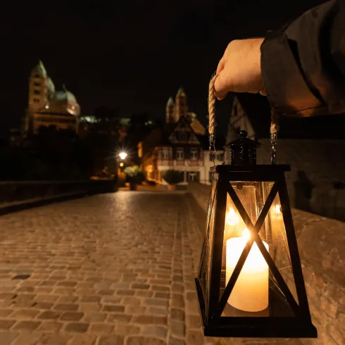 Eine Person hält eine Laterne auf einem Steinweg bei Nacht. Im Hintergrund sind historische Gebäude und eine Burg zu sehen, die sanft beleuchtet sind.