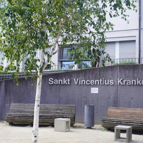 Ein Krankenhaus mit dem Namen "Sankt Vincentius Krankenhaus". Im Vordergrund befinden sich Holzbank und ein birkenähnlicher Baum.