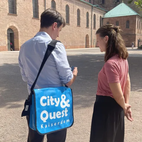 Zwei Personen stehen im Freien und sprechen miteinander. Eine Person trägt eine Tasche mit der Aufschrift „City & Quest Kaiserdom“.