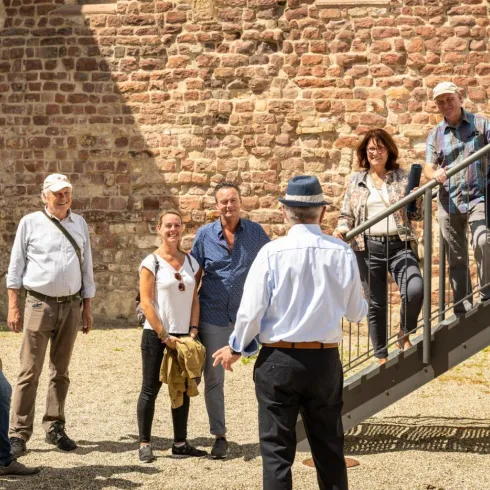 Eine Gruppe von Menschen hört einem Führer zu, der vor einer alten Mauer steht. Einige Teilnehmer stehen auf einer Treppe, während andere im Vordergrund stehen und aufmerksam zuhören.